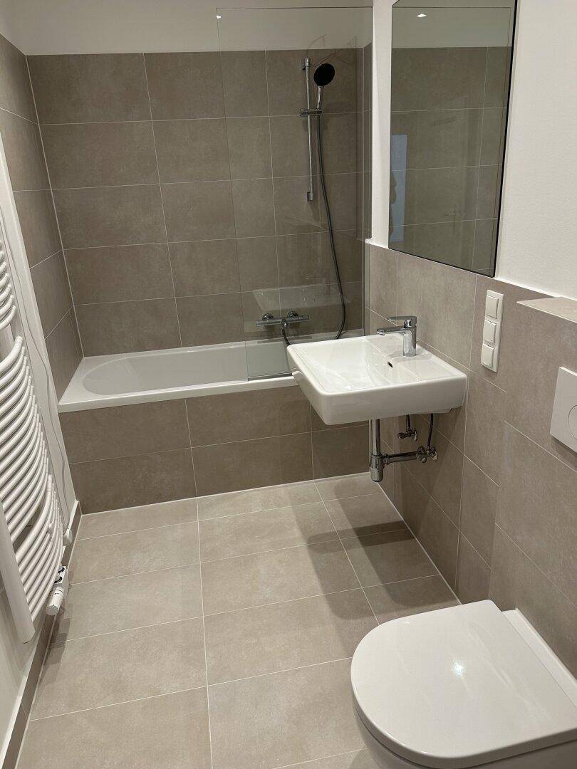 Modernes Badezimmer mit Badewanne, Waschbecken und WC, ausgestattet mit grauen Fliesen und Handtuchheizkörper.