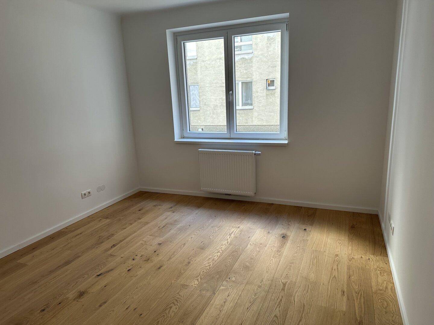 Geräumiges Zimmer mit Holzboden und großem Fenster, das viel Tageslicht hereinlässt und einen Blick auf den Innenhof bietet.