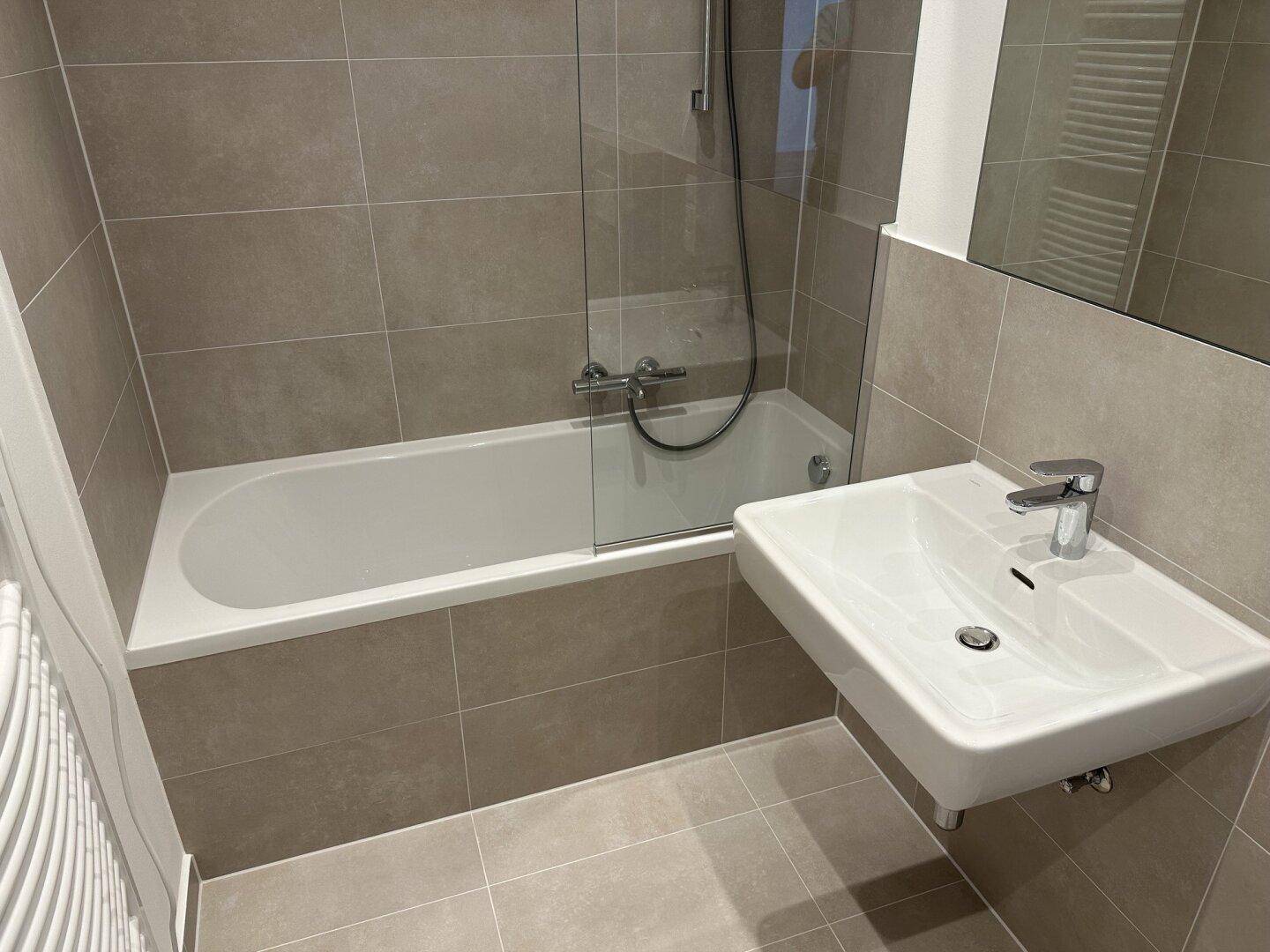 Modernes Badezimmer mit Badewanne, Duschvorrichtung und Waschbecken, ausgestattet mit grauen Fliesen.
