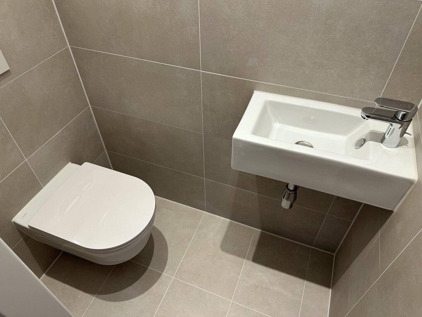 Separates WC mit wandhängender Toilette und kleinem Waschbecken, ausgestattet mit grauen Fliesen.