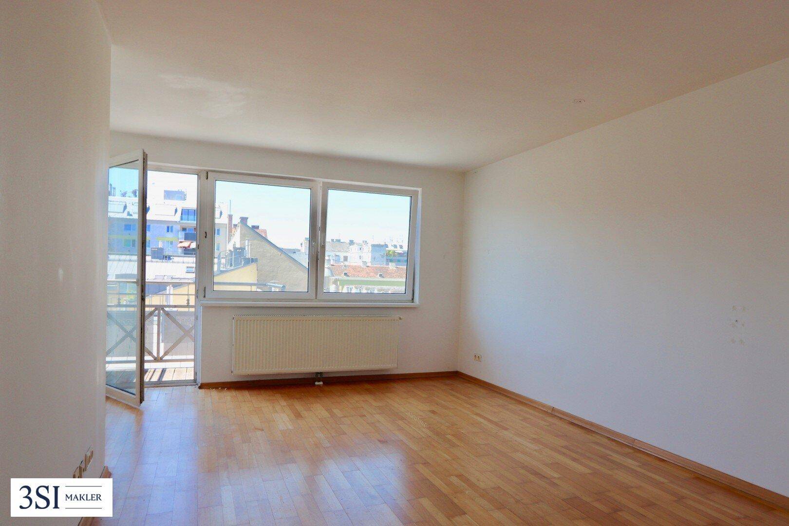 Helles, leeres Zimmer mit Holzfußboden, weißen Wänden und Zugang zu einem kleinen Balkon.
