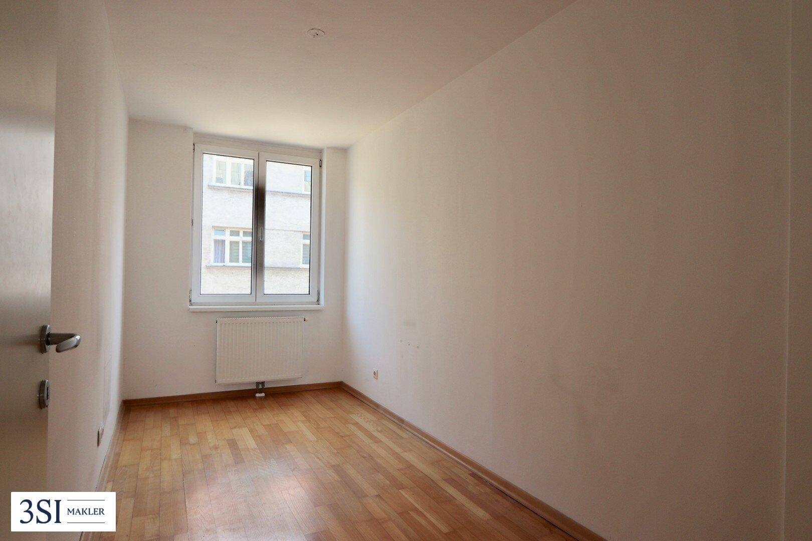 Leeres Schlafzimmer mit Holzparkettboden und einem Fenster, das viel Tageslicht hereinlässt.