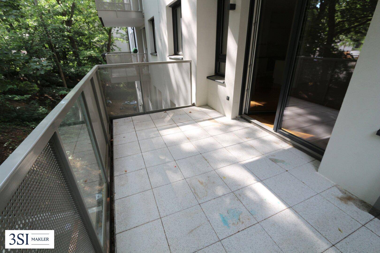 Geräumiger Balkon mit Fliesenboden und Glasgeländer, umgeben von Bäumen, bietet einen grünen Ausblick.