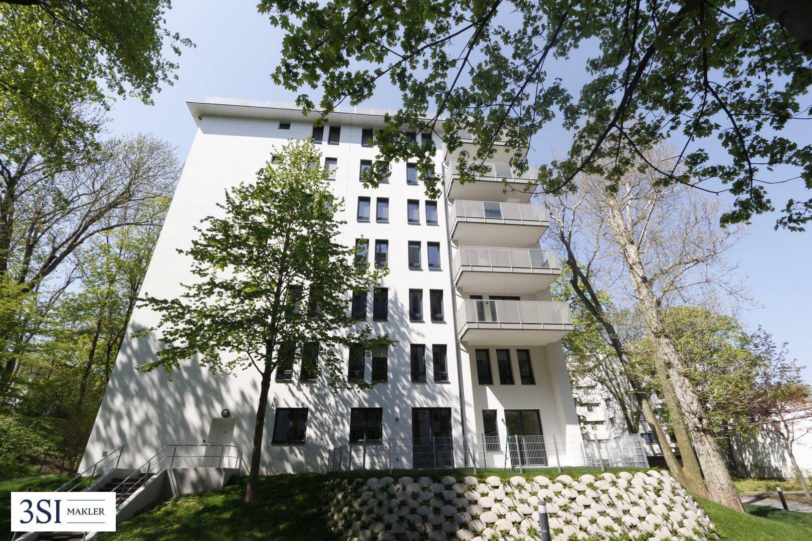 Modernes weißes Mehrfamilienhaus mit mehreren Balkonen, eingebettet in eine grüne Umgebung mit Bäumen.