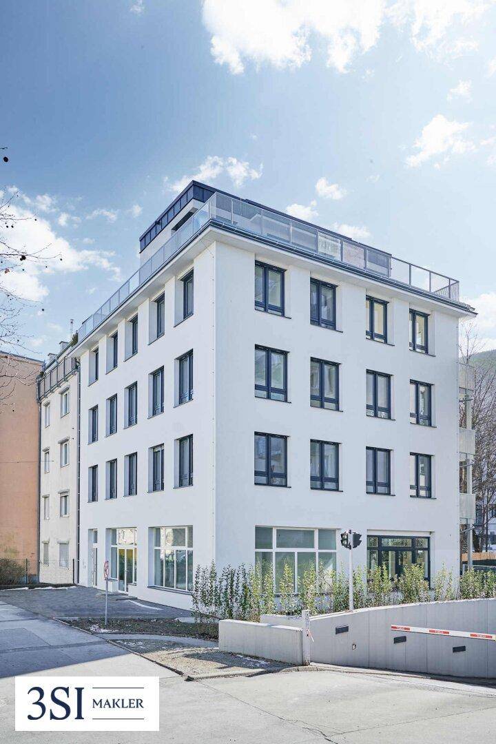 Modernes weißes Gebäude mit großen Fenstern, einer Dachterrasse und Zufahrt zur Tiefgarage.