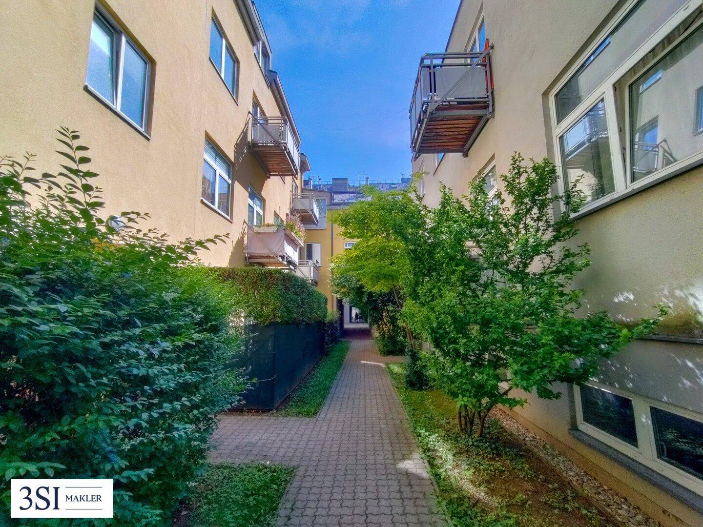 Schmaler Weg zwischen zwei Wohngebäuden mit grünen Hecken und kleinen Bäumen.