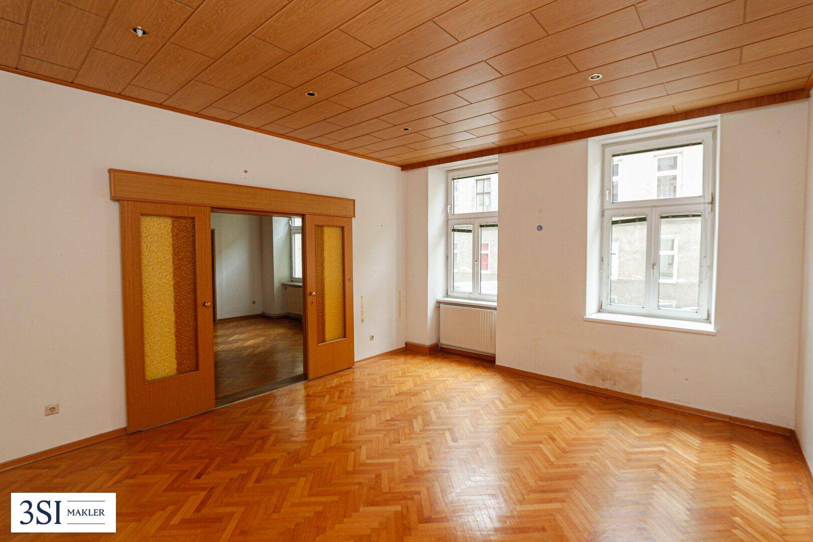 Helles Wohnzimmer mit zwei großen Fenstern, Fischgrätparkett und Schiebetüren zu einem weiteren Raum.