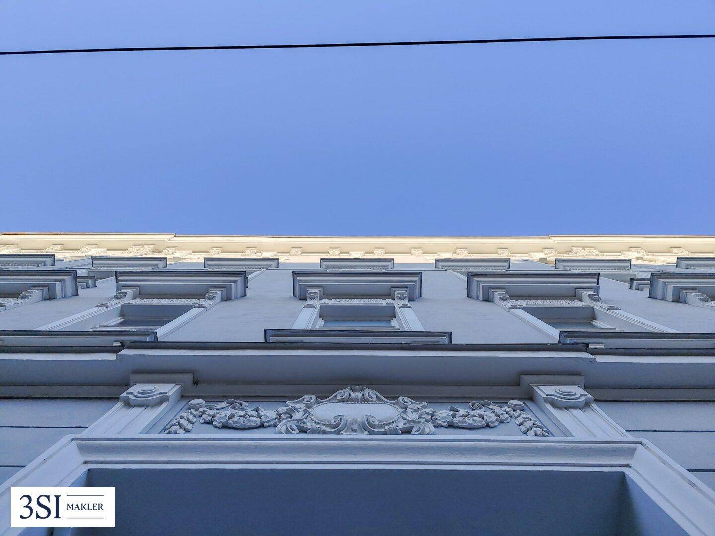 Detailaufnahme der historischen Fassade mit aufwendigen Stuckverzierungen und Fenstern unter blauem Himmel.