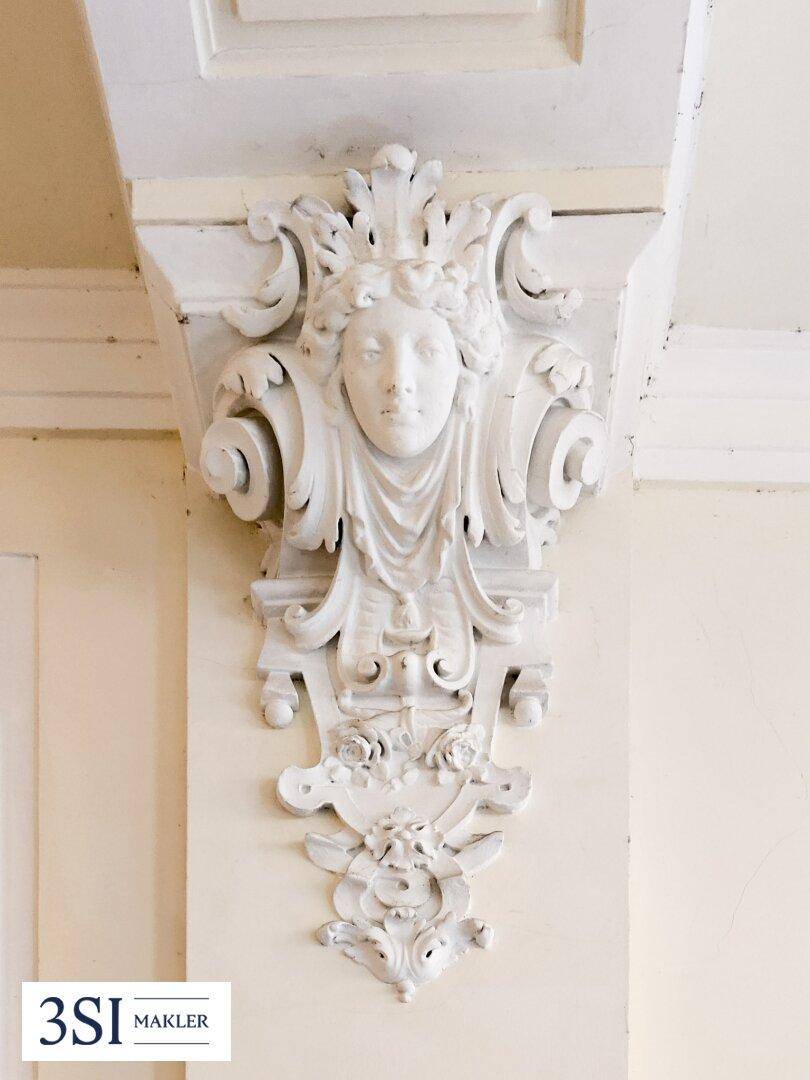Nahaufnahme einer detailreichen Stuckverzierung mit figürlichem Motiv an der Wand des historischen Flurs.
