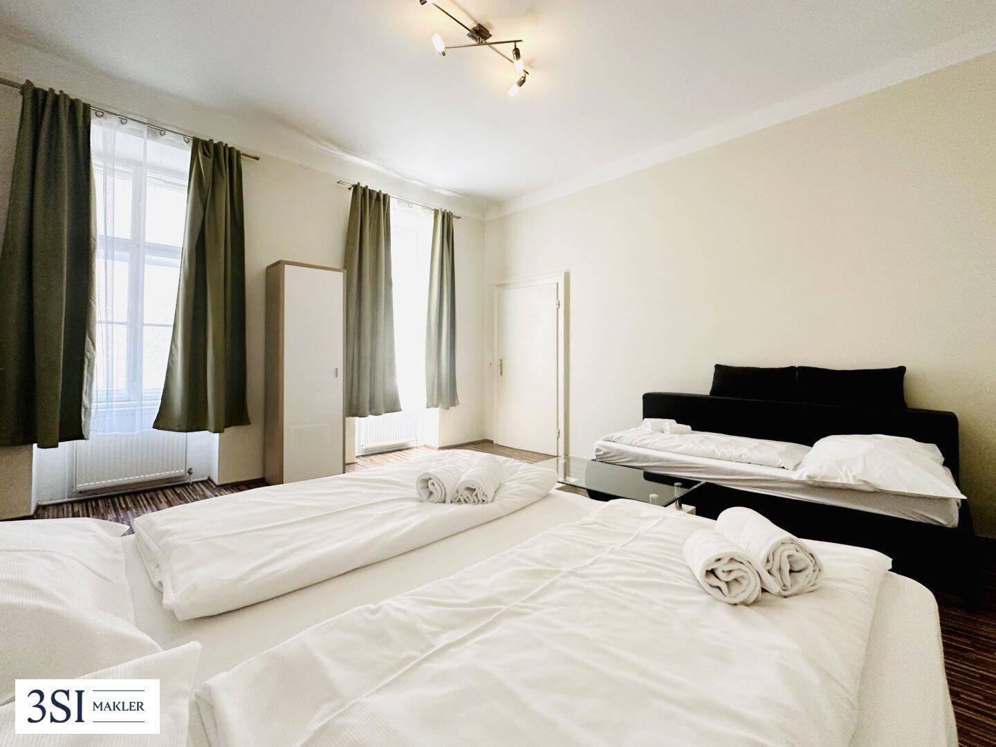 Helles Schlafzimmer mit zwei Betten, Parkettboden und hohen Fenstern mit Vorhängen.