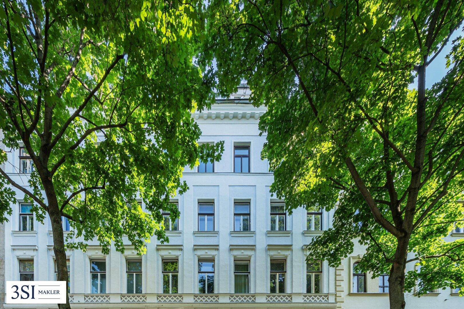 Elegante Gebäudefassade mit vielen Fenstern, umgeben von üppigem Grün und Sonnenlicht.