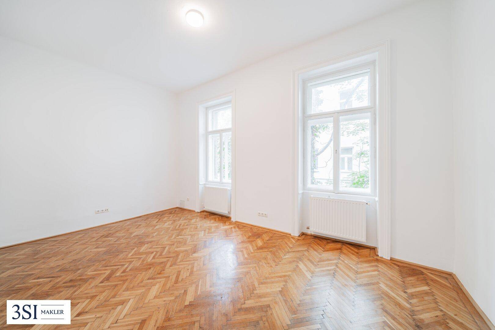 Geräumiges Zimmer mit Fischgrätparkett, hohen Fenstern und viel Tageslicht.