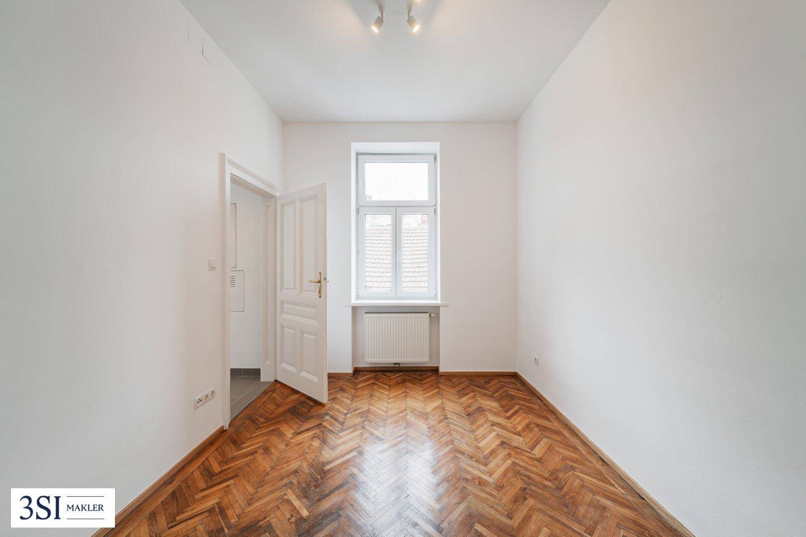 Kleines, helles Zimmer mit Fischgrätparkett und einem Fenster mit Blick ins Grüne.