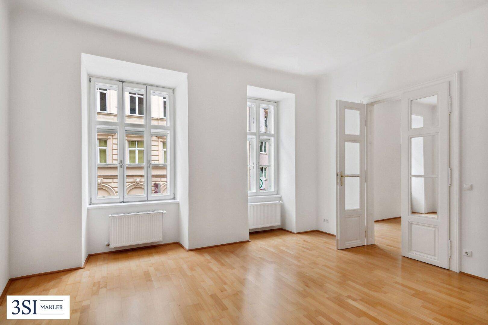 Helles Zimmer mit klassischem Parkettboden, hohen Fenstern und Heizkörpern unter den Fenstern.