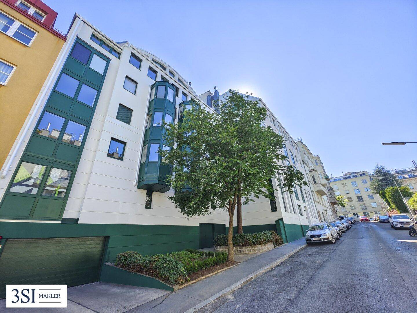 Moderne Wohnanlage an einer Straße mit Bäumen und geparkten Autos, helle Fassade.
