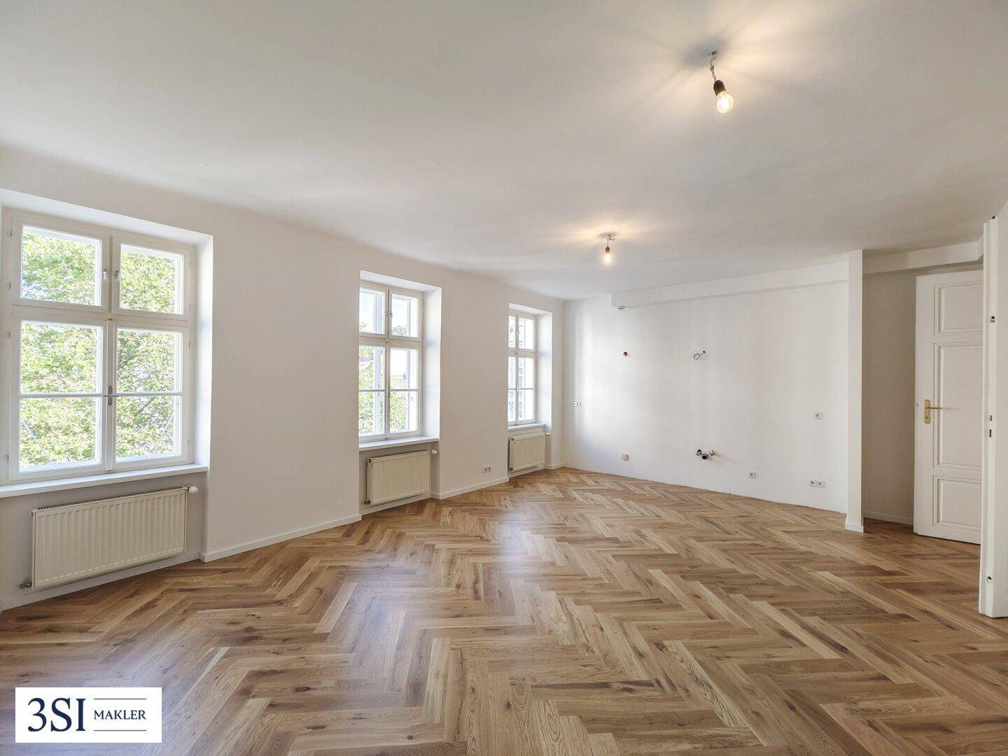 Geräumiges Zimmer mit elegantem Fischgrätparkett, großen Fenstern und viel Tageslicht.