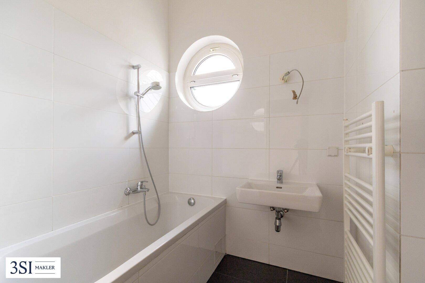 Helles Badezimmer mit Badewanne, Waschbecken und rundem Fenster, komplett gefliest.