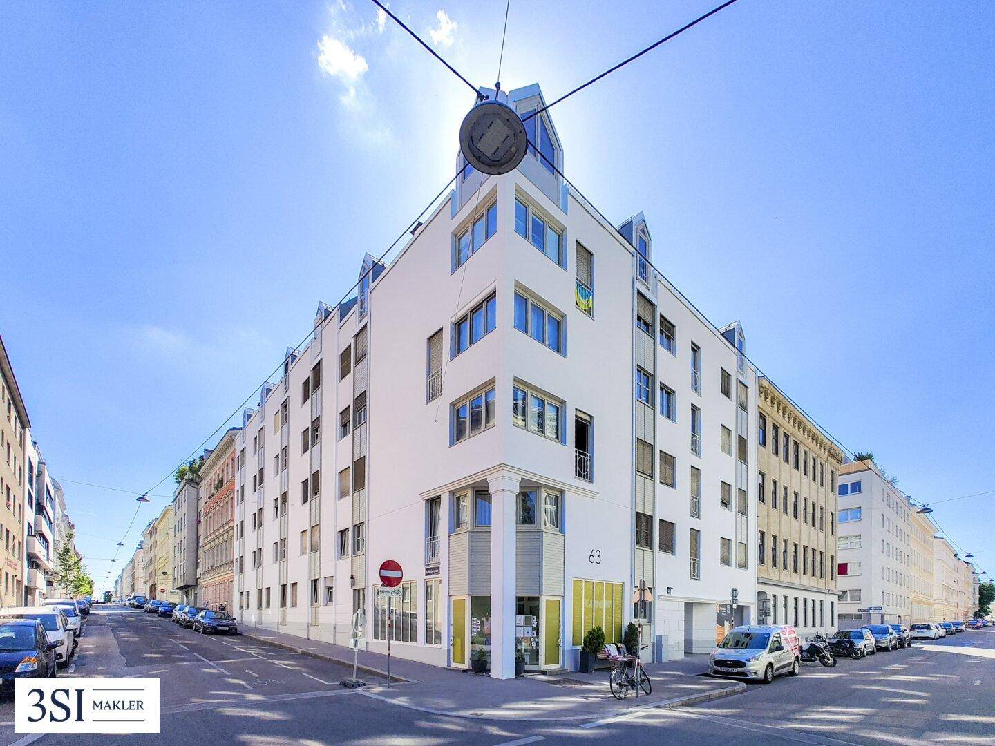 Modernes Eckgebäude mit weißer Fassade und vielen Fenstern an einer belebten Straße unter blauem Himmel.