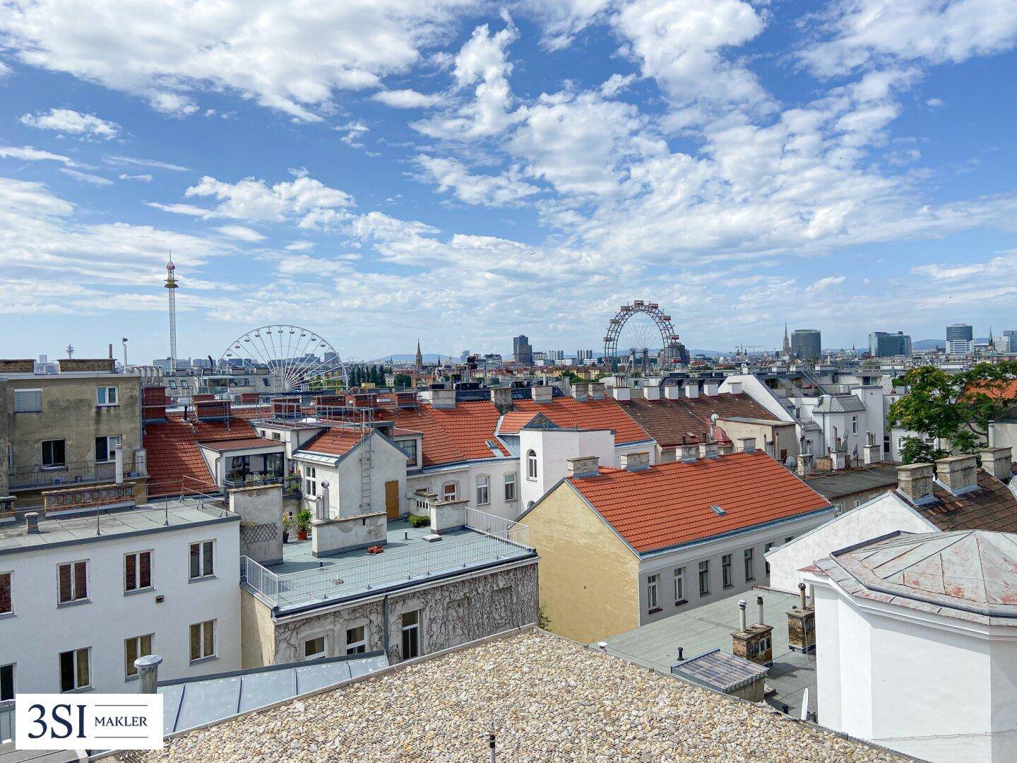 Weitläufiger Stadtblick über Dächer mit dem Riesenrad im Hintergrund unter blauem Himmel.