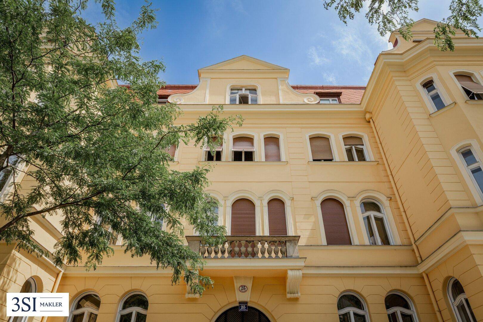 Gelbe Fassade eines Altbaus mit dekorativem Balkon und grünen Bäumen im Vordergrund.
