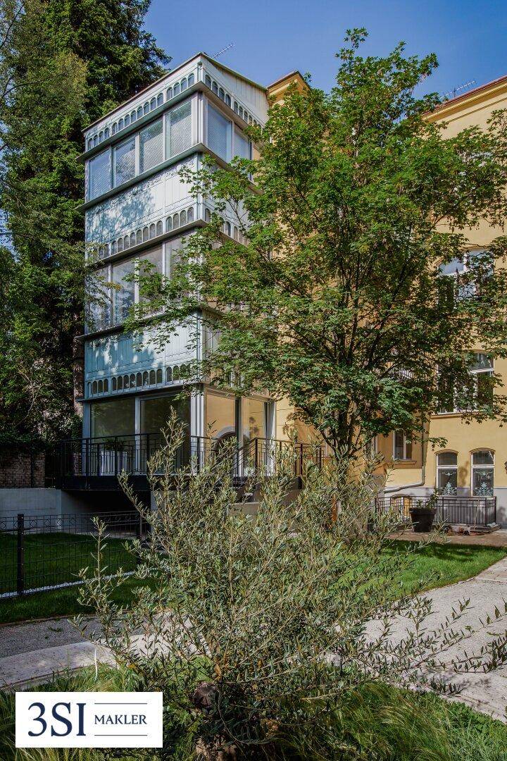 Seitenansicht eines Gebäudes mit modernen Glasanbauten und umgebenden Bäumen.