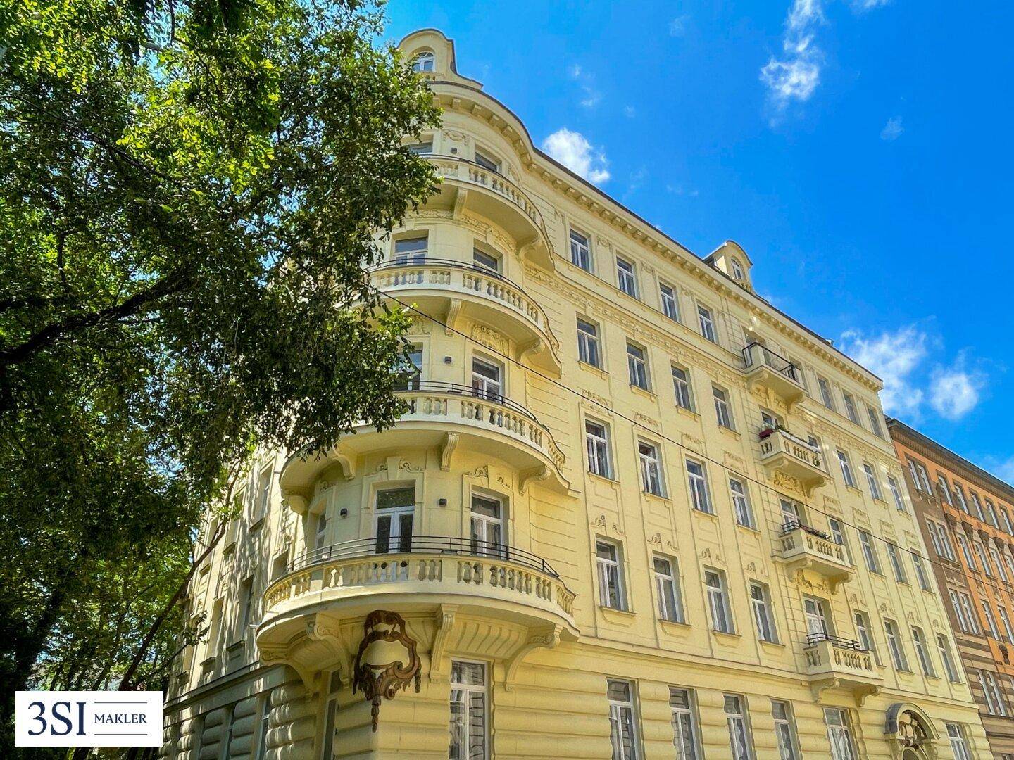 Historisches Gebäude mit reich verzierter Fassade, Balkonen und grünen Bäumen im Vordergrund.