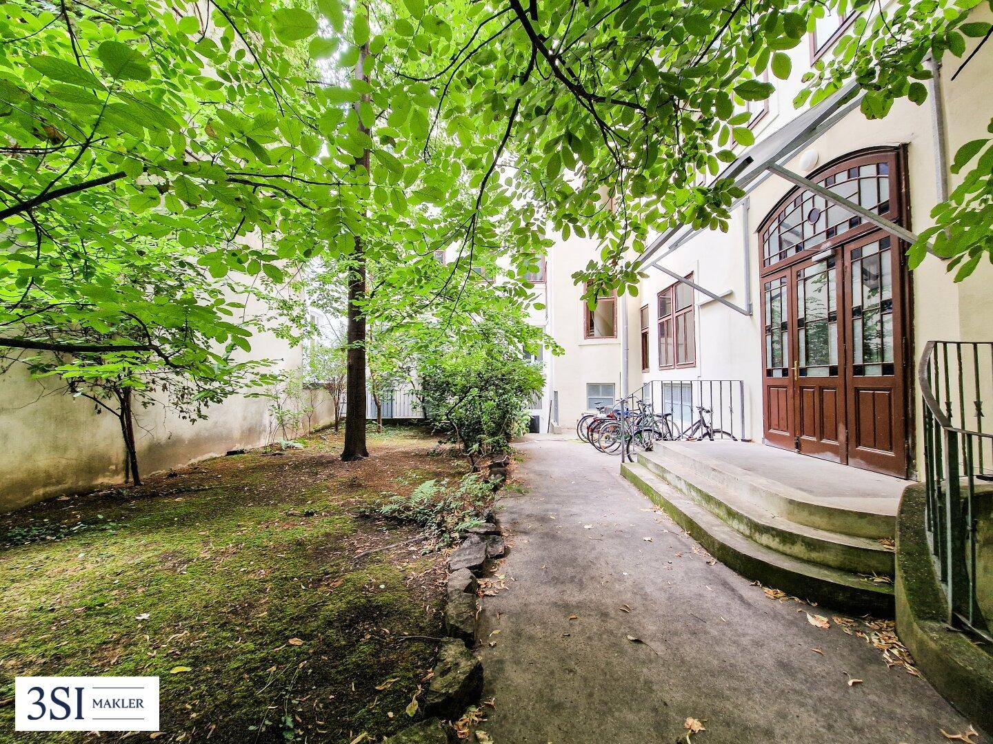 Grüner Innenhof mit altem Baumbestand, Rasenfläche und einem Eingang zu den Wohnungen.