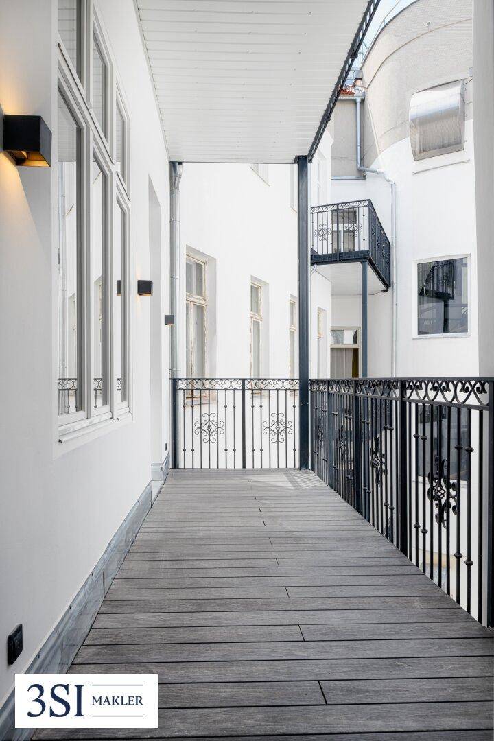 Langer, schmaler Balkon mit Holzboden und schmiedeeisernem Geländer, umgeben von Gebäuden.