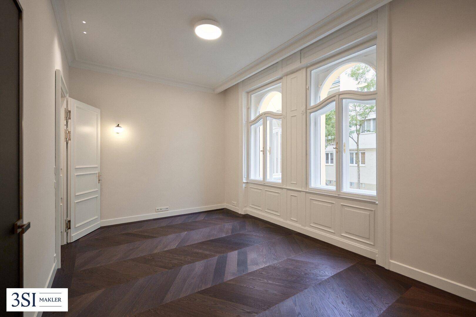 Leerer Raum mit elegantem Fischgrätparkett und hohen, stilvollen Fenstern, die viel Licht spenden.