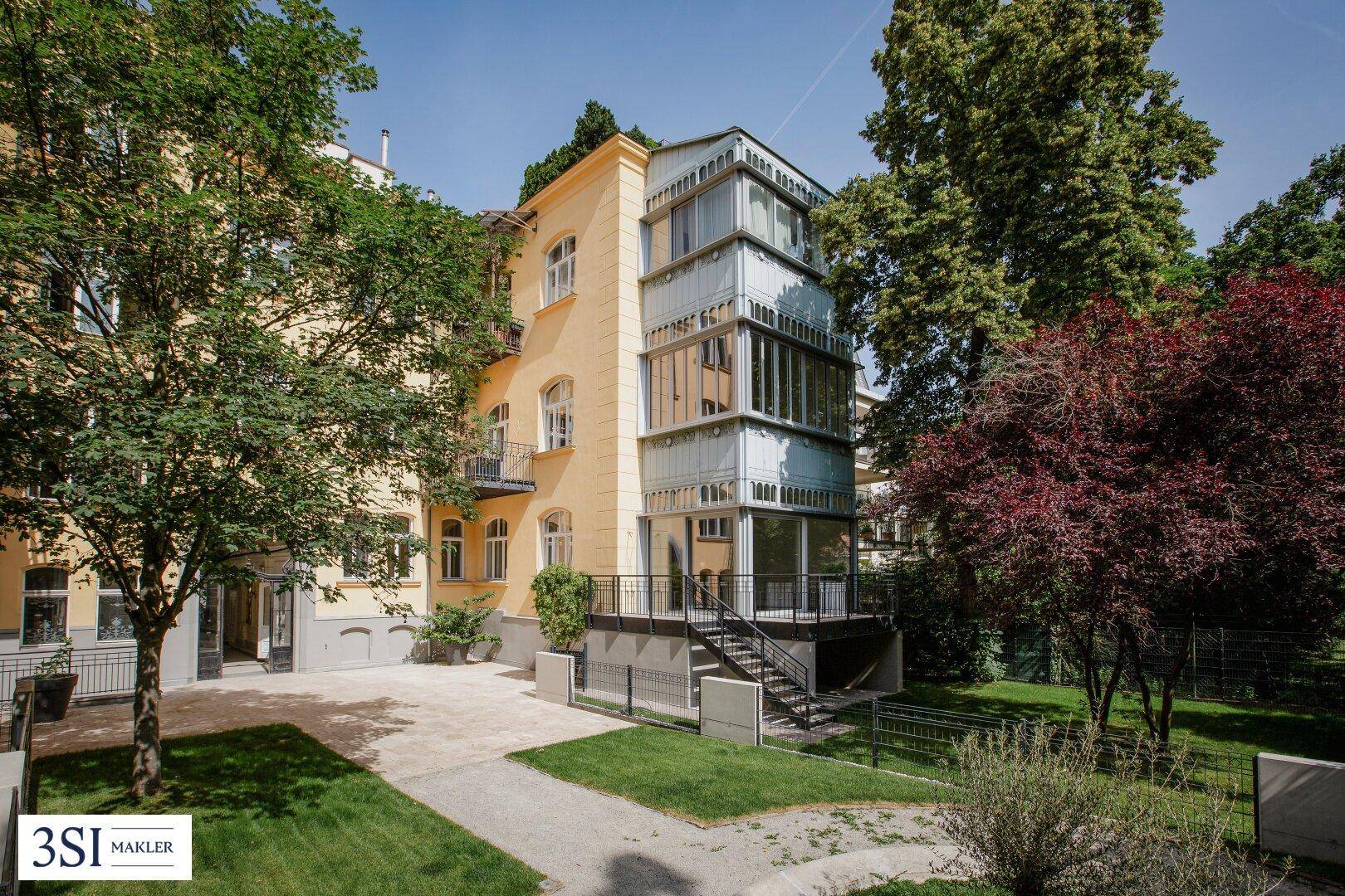 Historisches Gebäude mit gelber Fassade und modernem Glasanbau, umgeben von altem Baumbestand.
