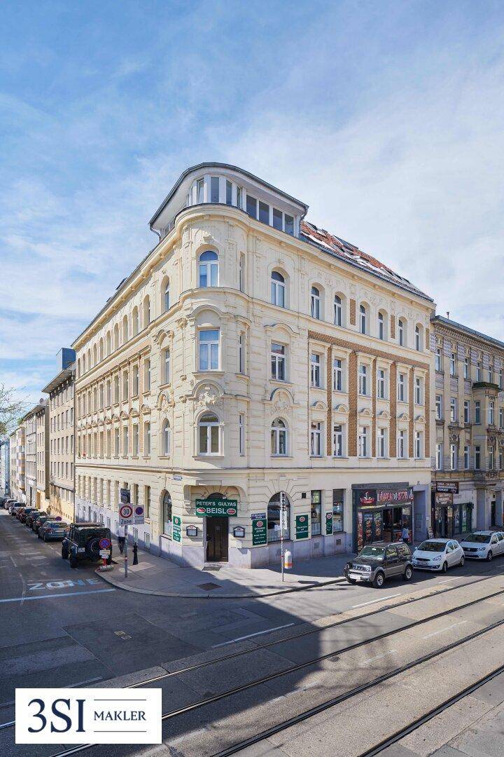 Historisches Eckgebäude mit Geschäften im Erdgeschoss und Straßenbahnschienen im Vordergrund.