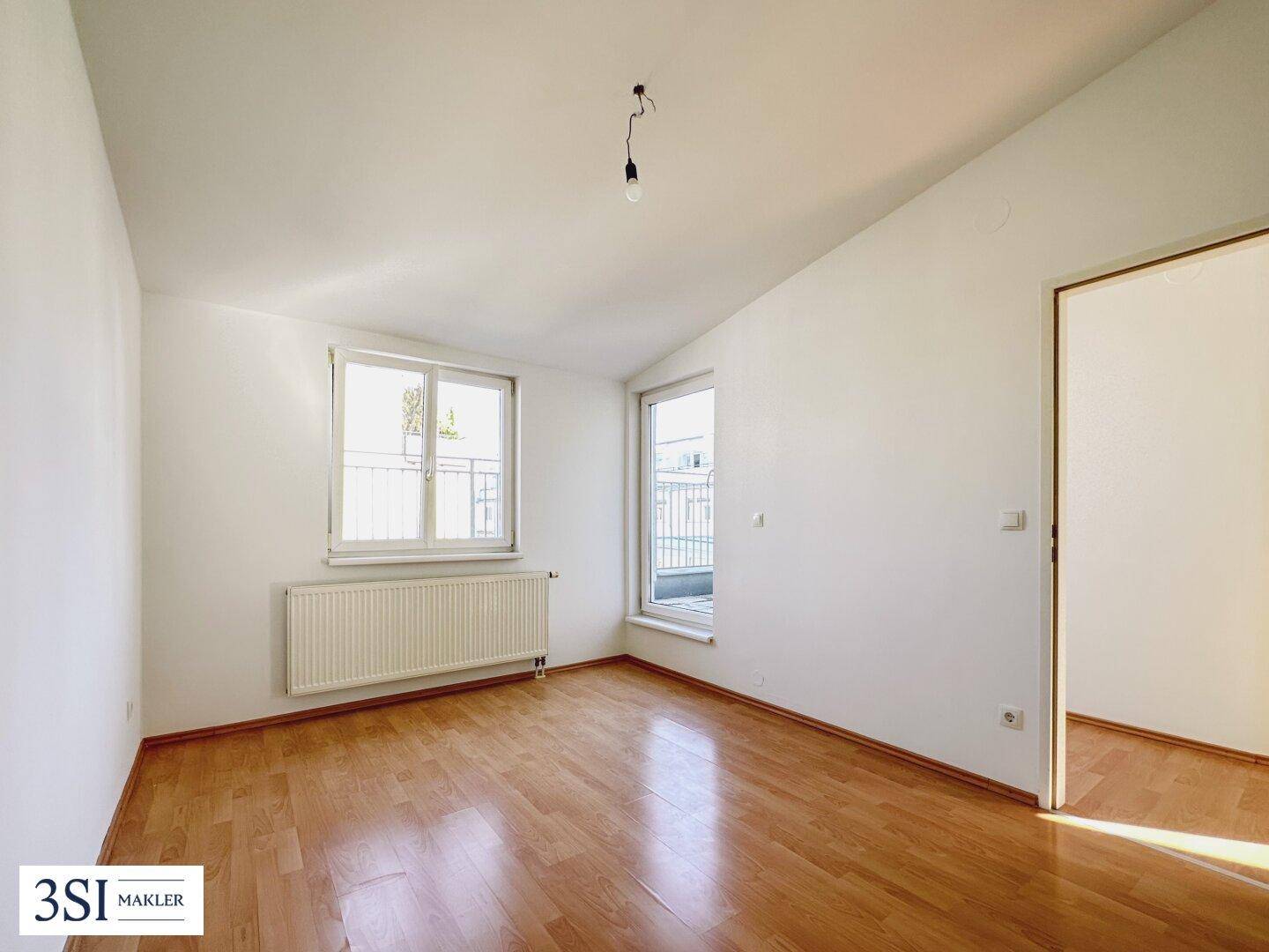 Leeres Zimmer mit Holzboden, Heizkörper und Zugang zu einem französischen Balkon, ideal als Schlafzimmer.