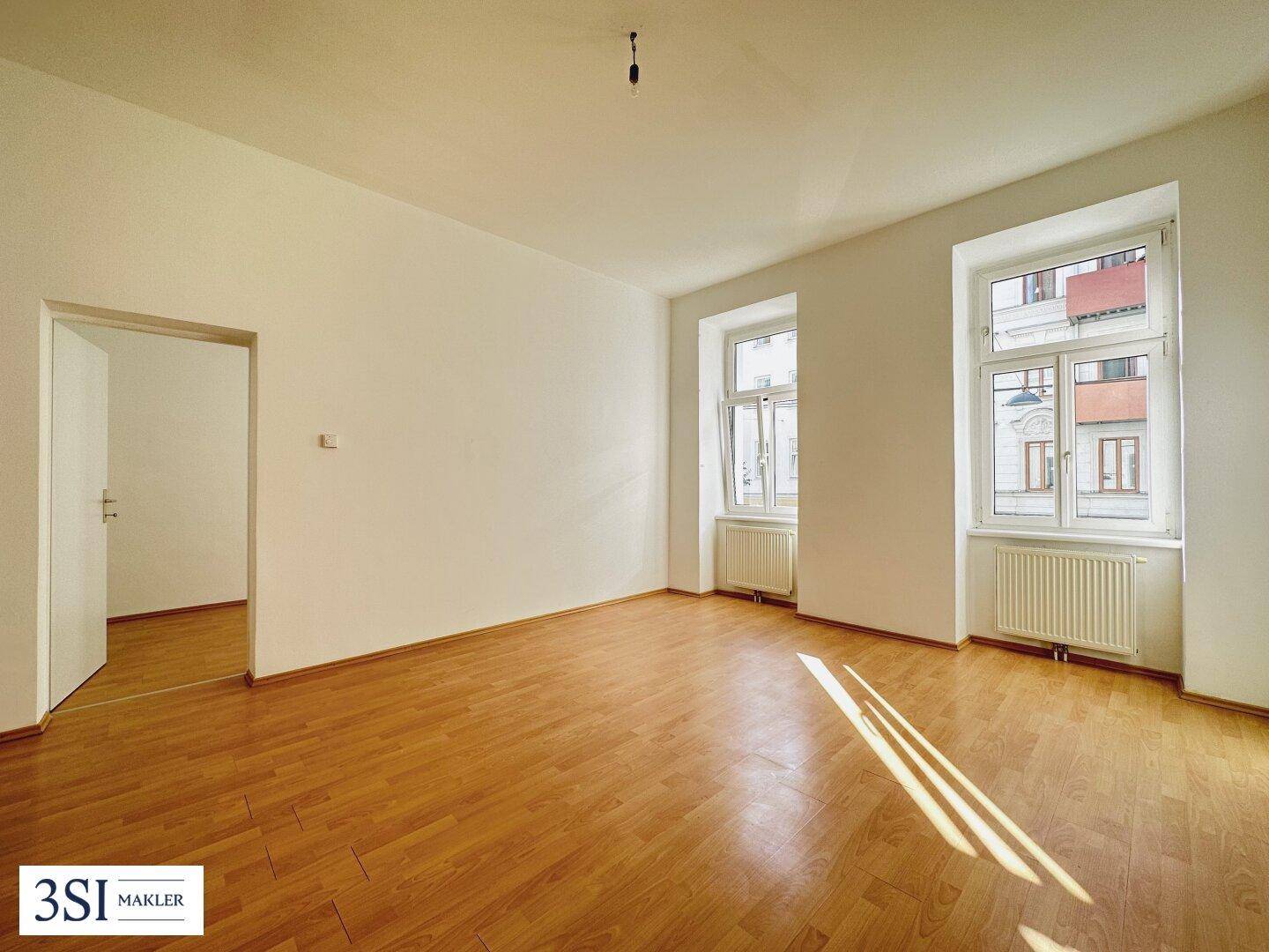 Geräumiges, leeres Wohnzimmer mit Holzboden und zwei großen Fenstern, die viel Licht hereinlassen.