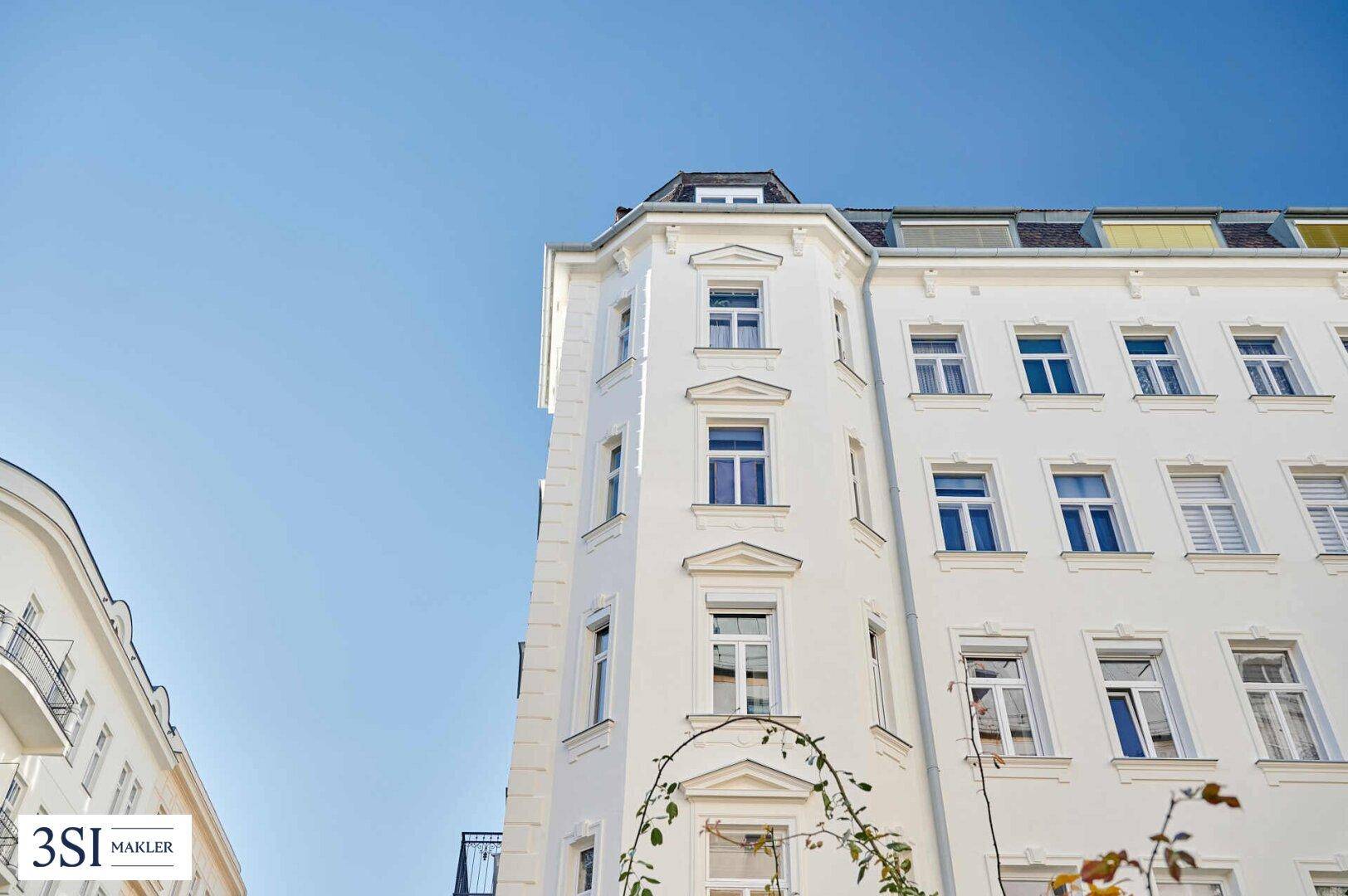 Helle Fassade eines Eckgebäudes unter blauem Himmel mit klassischen architektonischen Details.