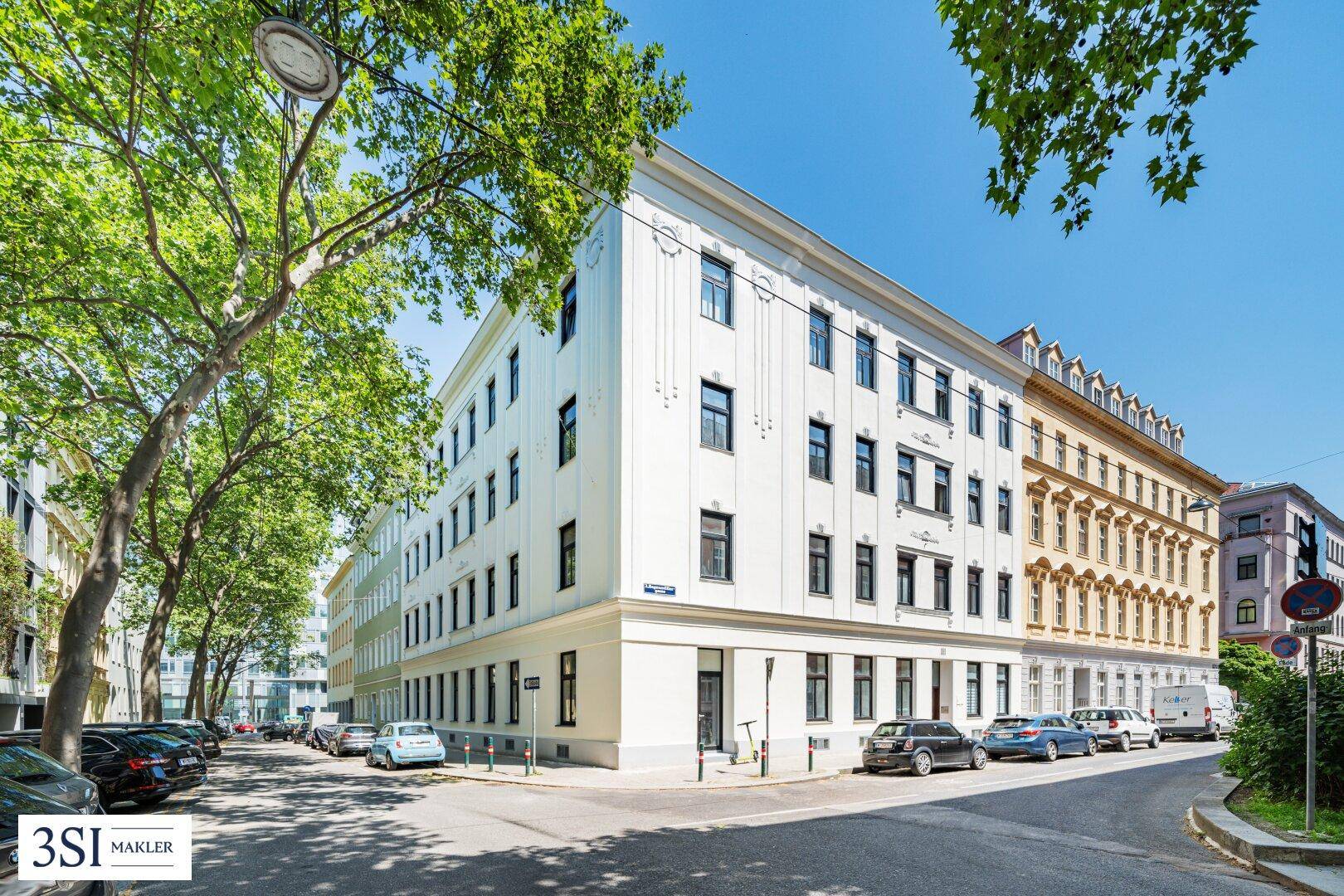 Helles Eckgebäude mit klassischer Fassade an einer belebten Straße unter blauem Himmel.