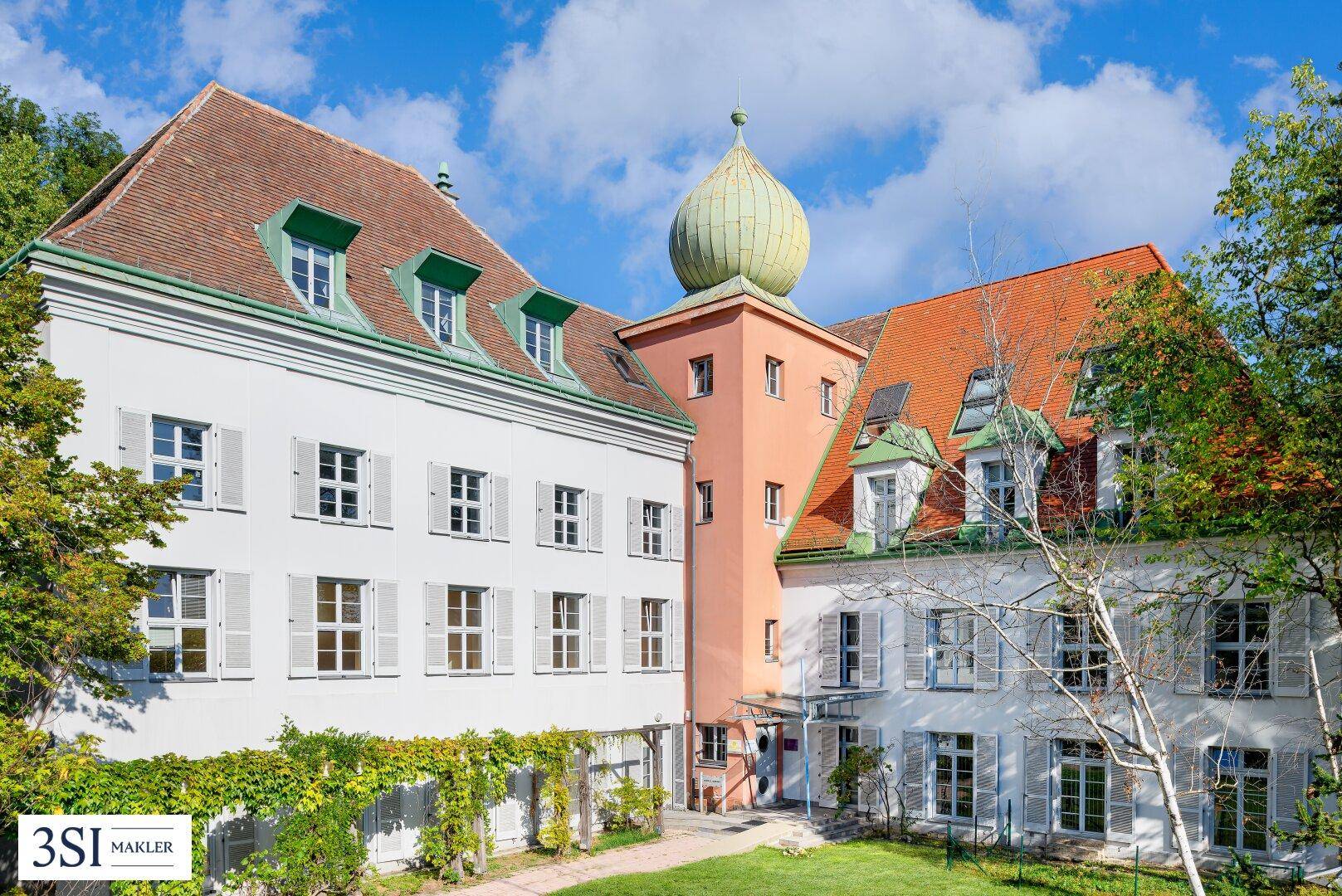 Historisches Gebäude mit markantem Turm, roten Dächern und vielen Fenstern in einer grünen Umgebung.