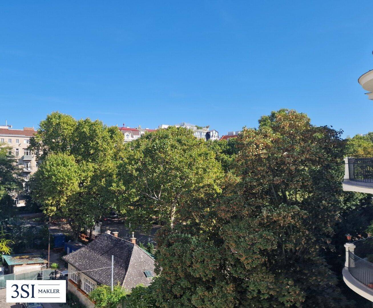 Grüne Aussicht von einem Balkon mit vielen Bäumen und einigen Dächern unter klarem, blauem Himmel.
