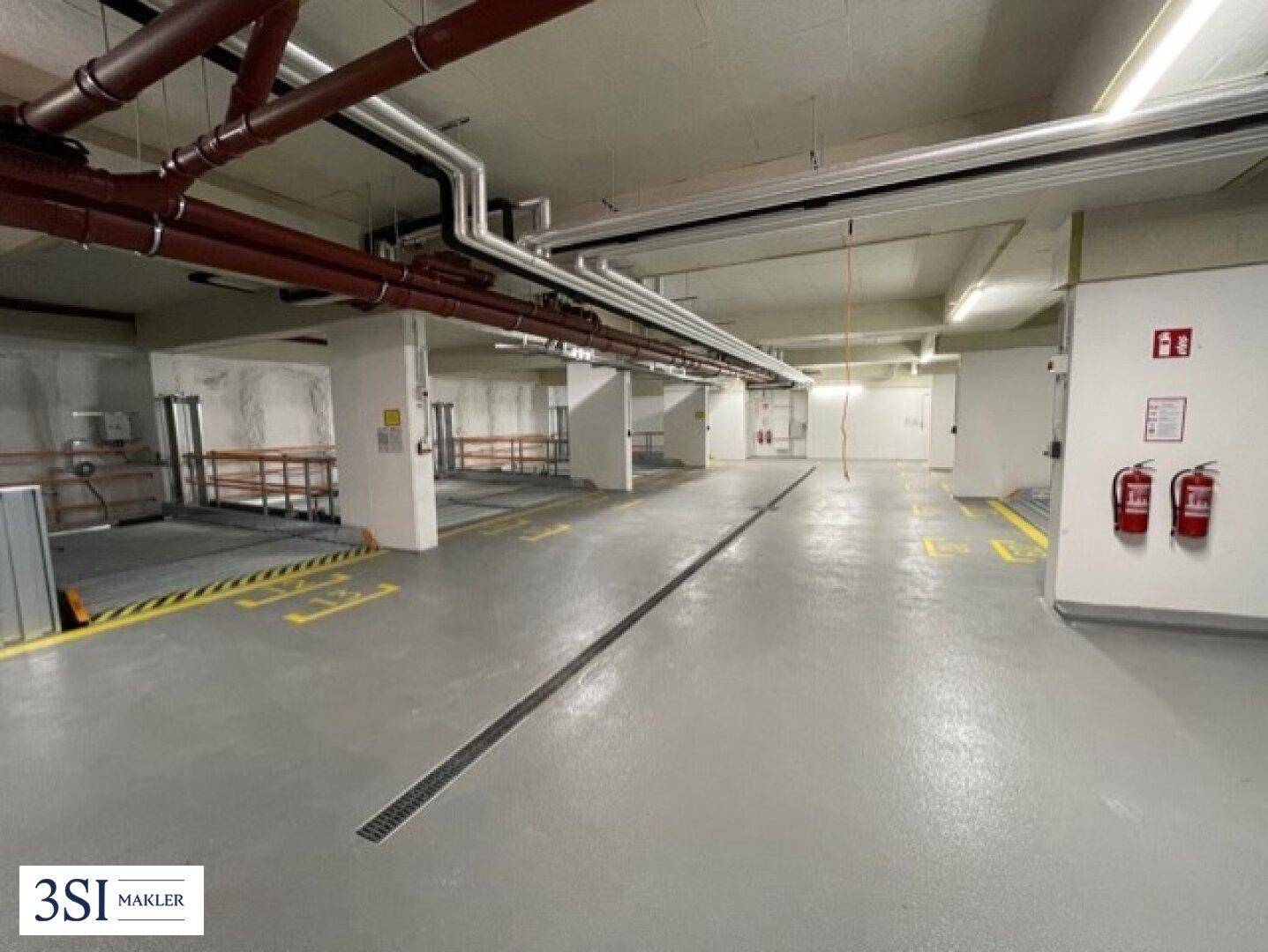 Weitläufige Tiefgarage mit Parksystemen, sichtbaren Leitungen und Brandschutzausrüstung.