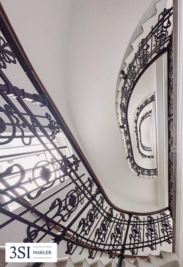 Blick nach oben in ein elegantes Treppenhaus mit kunstvollem schmiedeeisernem Geländer.