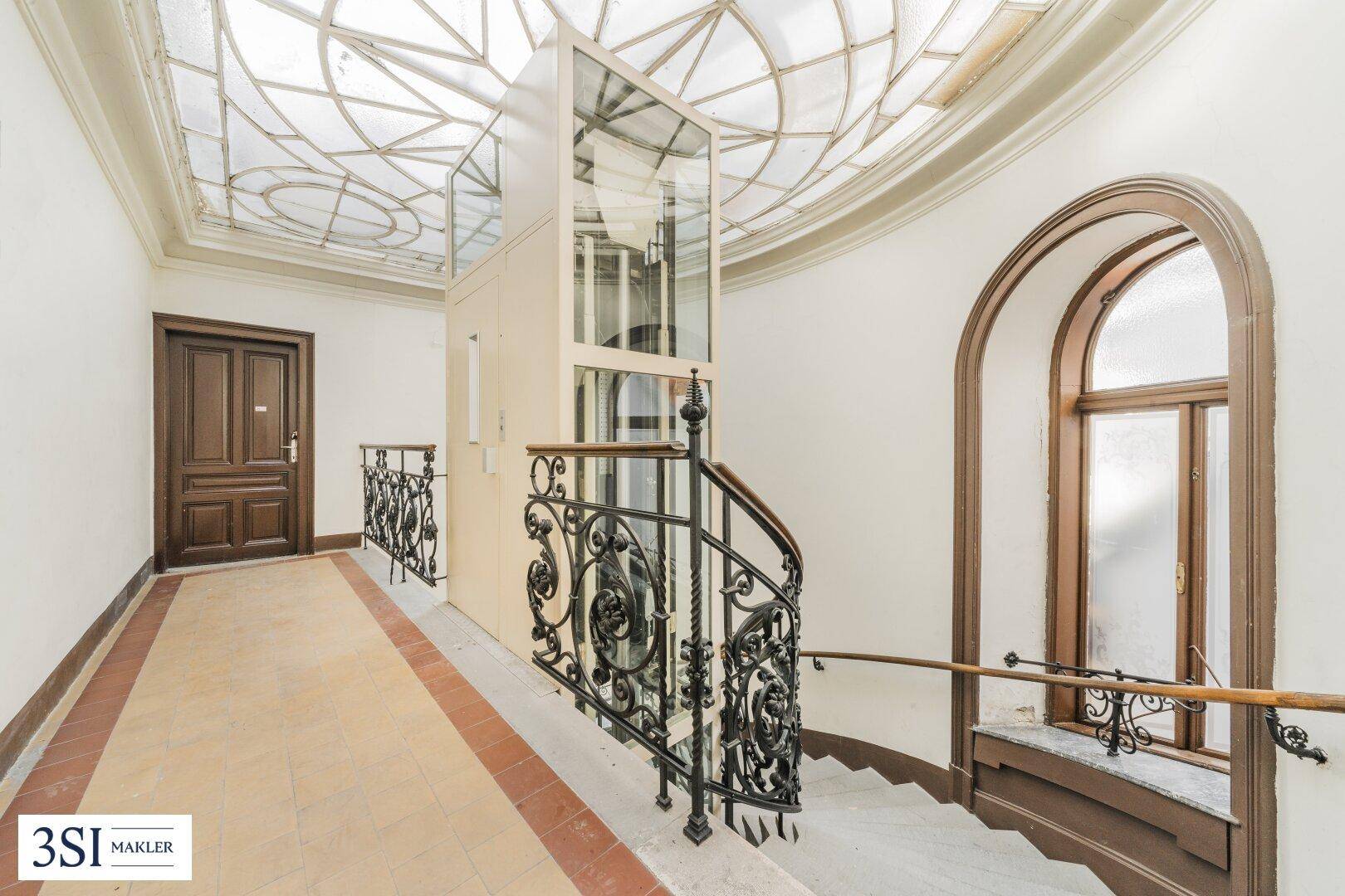 Historisches Treppenhaus mit schmiedeeisernem Geländer und einem modernen Glasaufzug.