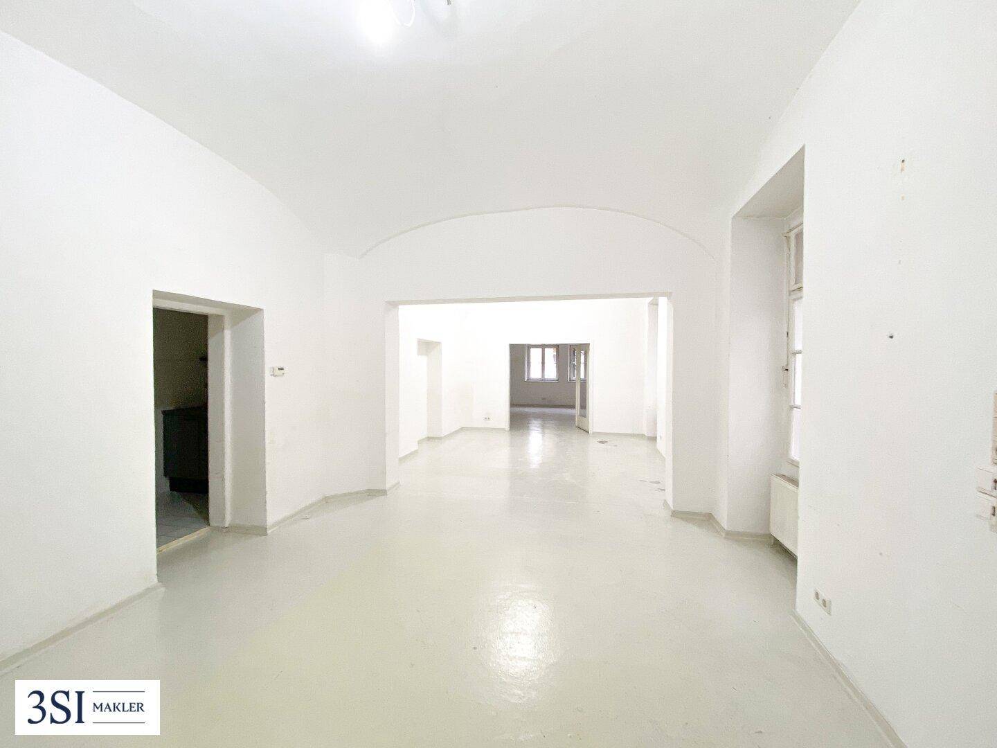 Großer, leerer Raum mit hellen Wänden, gewölbter Decke und offenem Durchgang zu weiteren Bereichen.
