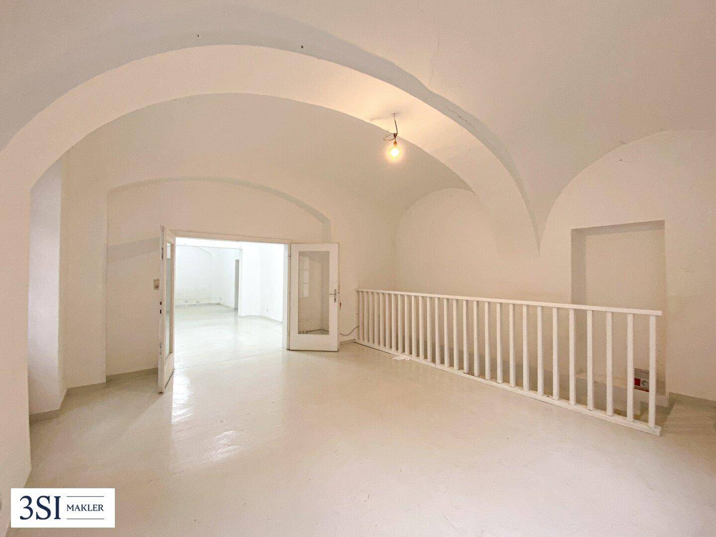Heller, leerer Raum mit gewölbter Decke, weißer Balustrade und offenem Zugang zu weiteren Bereichen.
