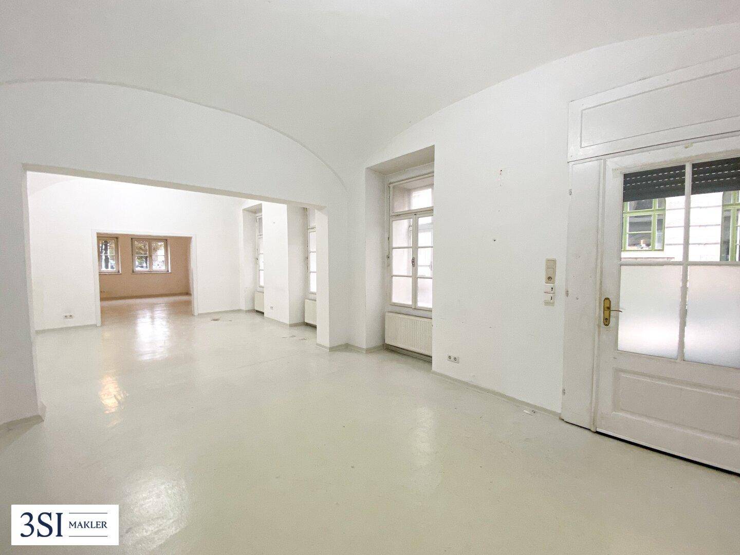 Heller, leerer Raum mit gewölbter Decke, großen Fenstern und einem Durchgang zu angrenzenden Zimmern.