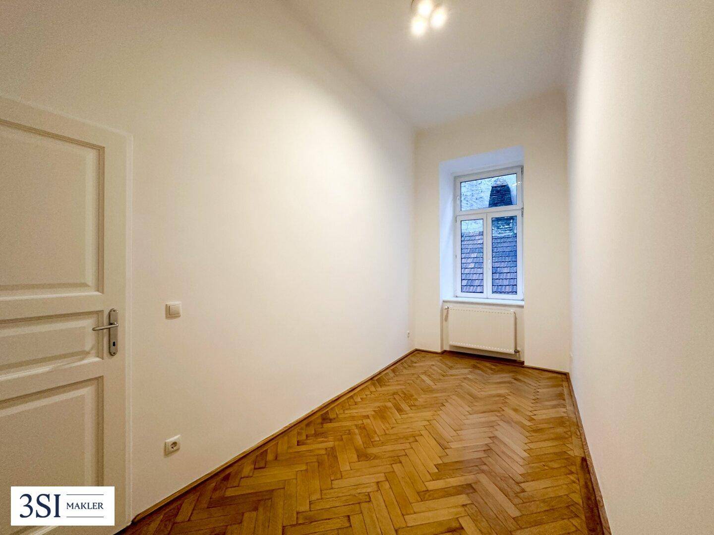 Leeres Zimmer mit klassischem Fischgrätparkett und einem Fenster für natürliche Belichtung.