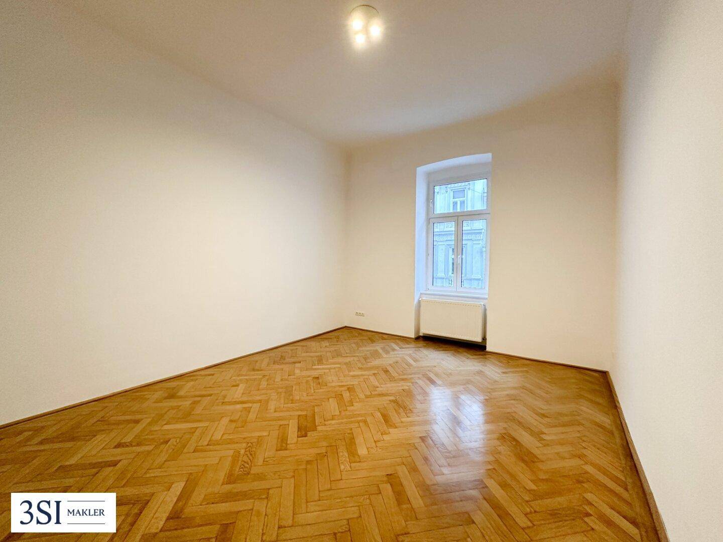 Helles Zimmer mit Fischgrätparkett und einem Fenster, das viel Tageslicht hereinlässt.