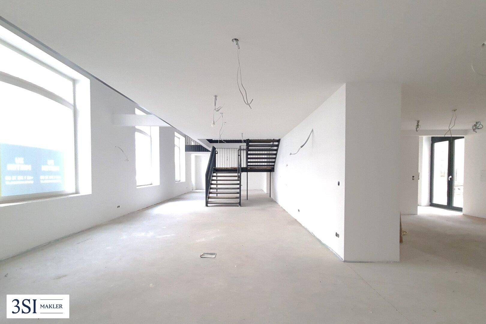 Weitläufiger, heller Innenraum im Rohbau mit großen Fenstern und einer modernen Treppenkonstruktion.