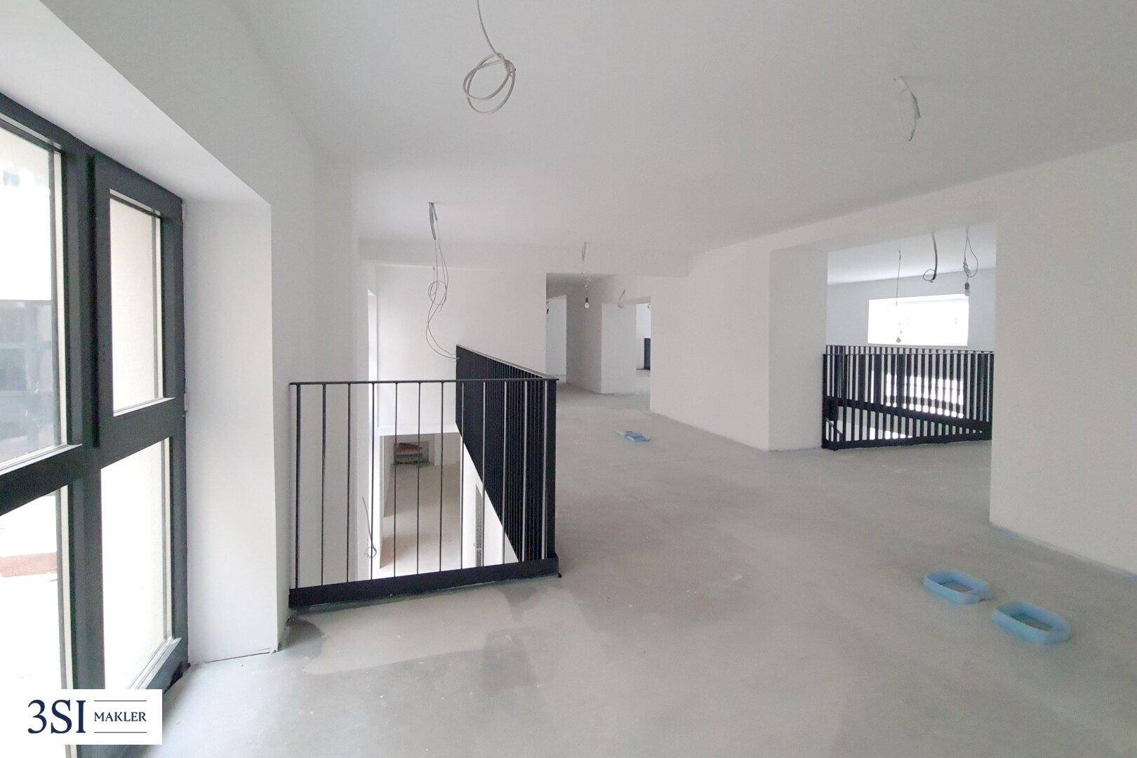 Unfertiger Innenraum mit hohen Decken, großen Fenstern und Betonboden, bereit für den Ausbau.