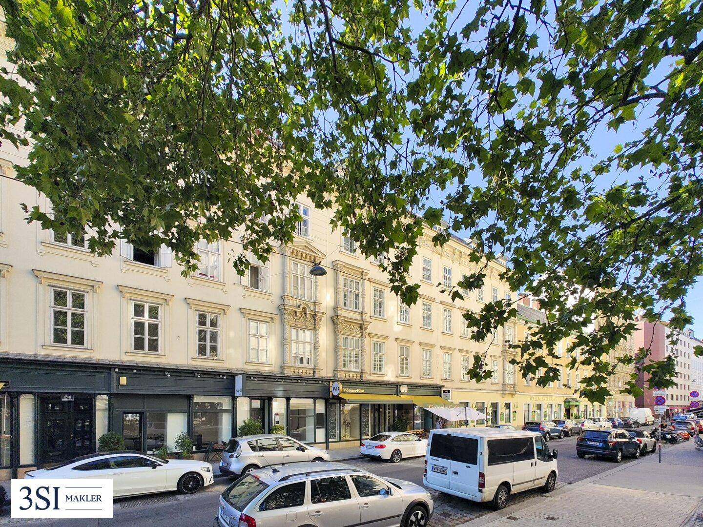 Historische Gebäudefassade an einer belebten Straße mit parkenden Autos und grünen Bäumen.