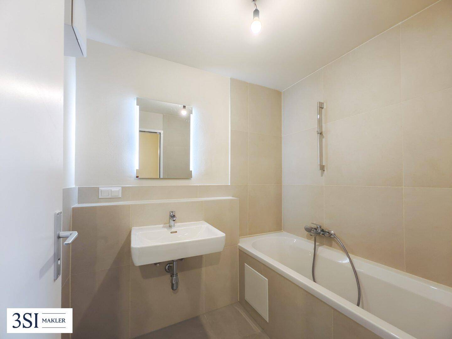 Modernes Badezimmer mit Badewanne, Einzelwaschbecken und beleuchtetem Spiegel in hellen Beigetönen.