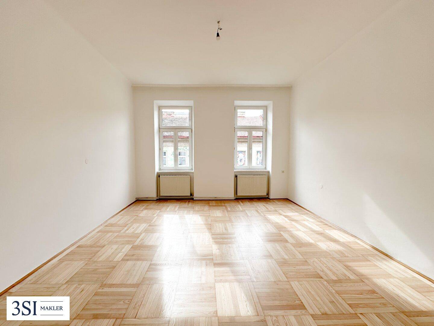 Helles, leeres Zimmer mit Fischgrätparkett und zwei großen Fenstern, die viel Tageslicht hereinlassen.